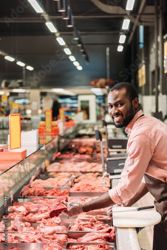 african american male butcher in apron holding steak of raw meat and looking at camera in supermarket
