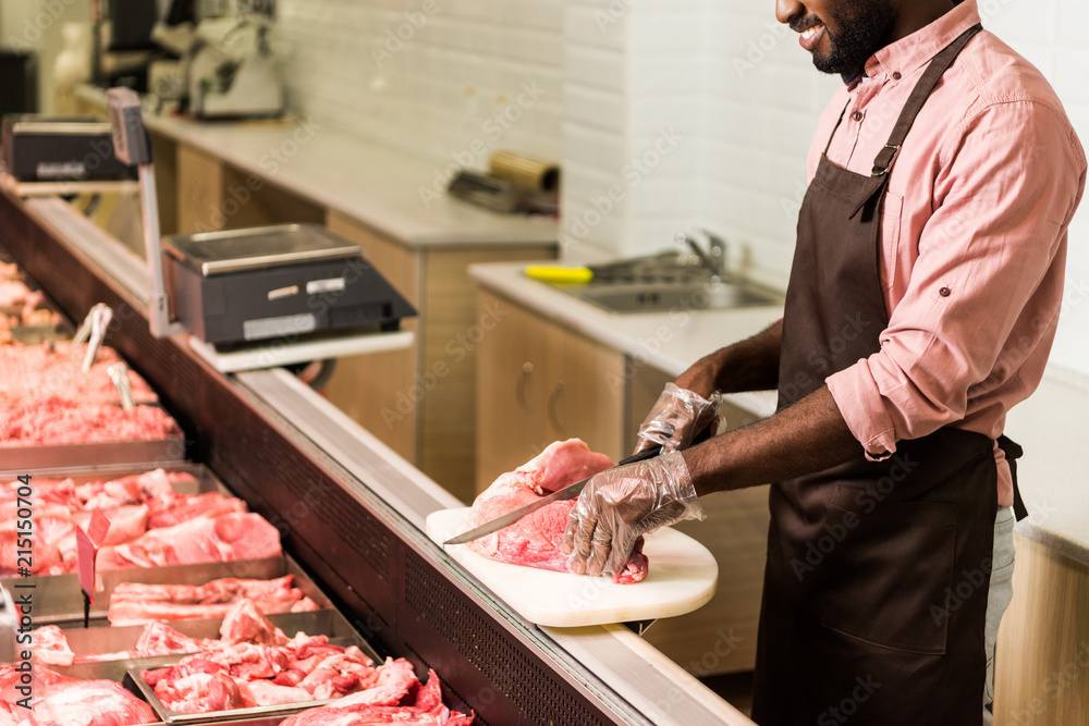 partial view of smiling african american male shop assistant in apron cutting steak of raw meat in hypermarket