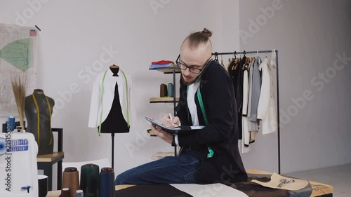 Young fashion designer or tailor sitting on table in atelier and talking phone and makes notes in his notebook. Young bearded man in glasses dressed in white shirt and black blouse with measure tape photo