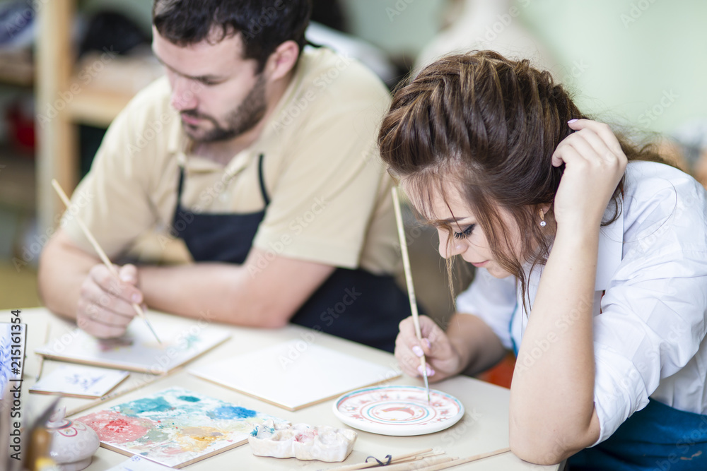 Two Young Caucasian Ceramists Painting and Glazing Clay Crafts Together in Workshop with Paintbrushes. Two Young Caucasian Ceramists Painting and Glazing Clay Crafts Together in Workshop with Paintbrushes.