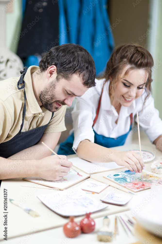 Naklejka premium Pottering Ideas. Caucasian Ceramists Painting and Glazing Clay Crafts Together in Workshop Using Paintbrushes. Woman Looking at camera.