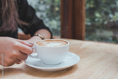 Closeup image of a woman holding a white cup of hot coffee on wooden table in cafe