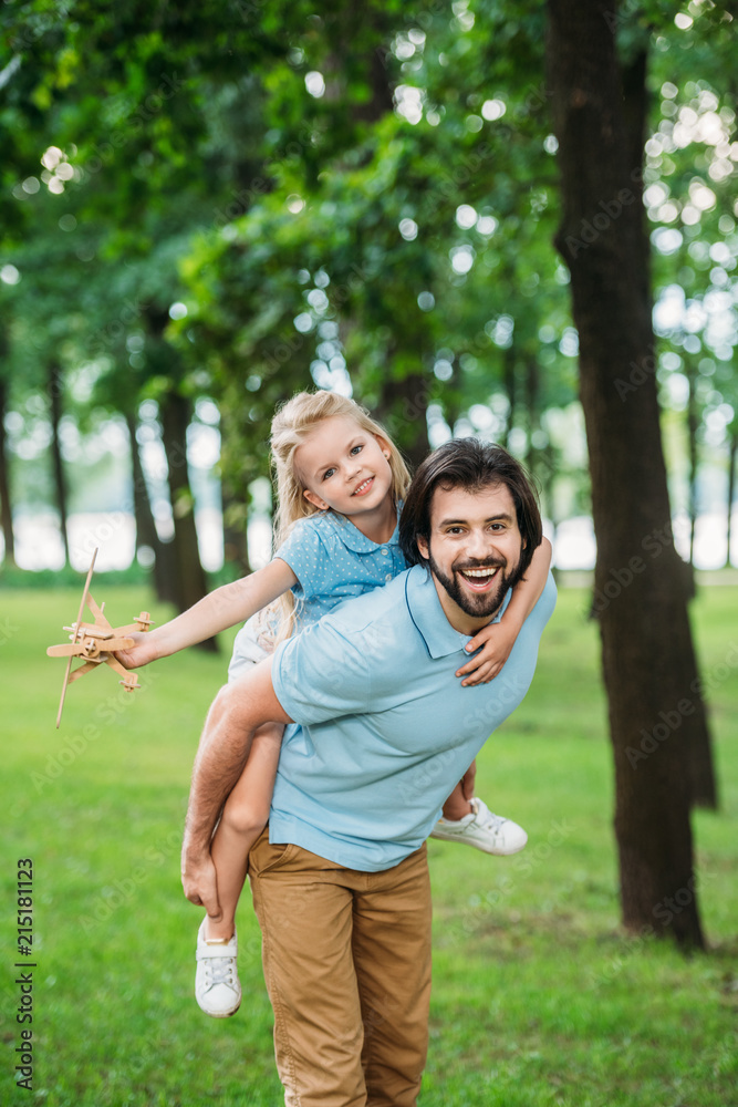 Fototapeta premium adorable daughter piggybacking on happy father and playing with toy plane at park