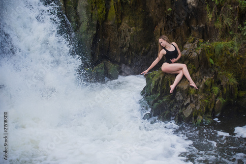 Young blond woman in a black swimwear sitting by the waterfall.  Young woman enjoying nature