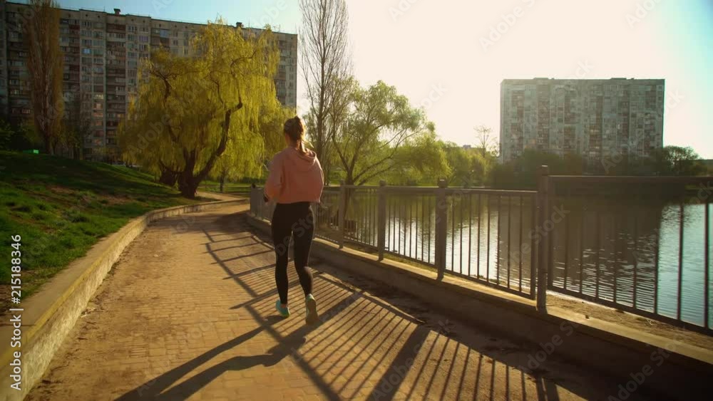 back view woman jogging outdoors. girl wearing sportswear black leggings and pink hoodie, jogger running around the lake in spring season back view woman jogging outdoors. girl wearing sportswear black leggings and pink hoodie, jogger running around the lake in spring season
