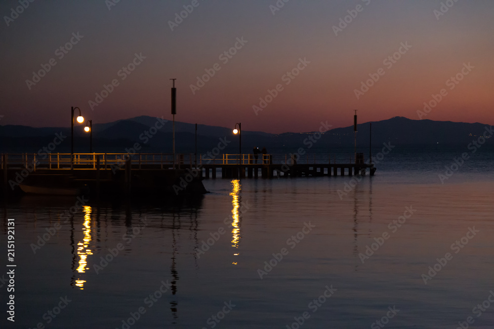 Some people on a pier at Trasimeno lake (Umbria, Italy) at dusk, with beautiful water reflections and warm colors