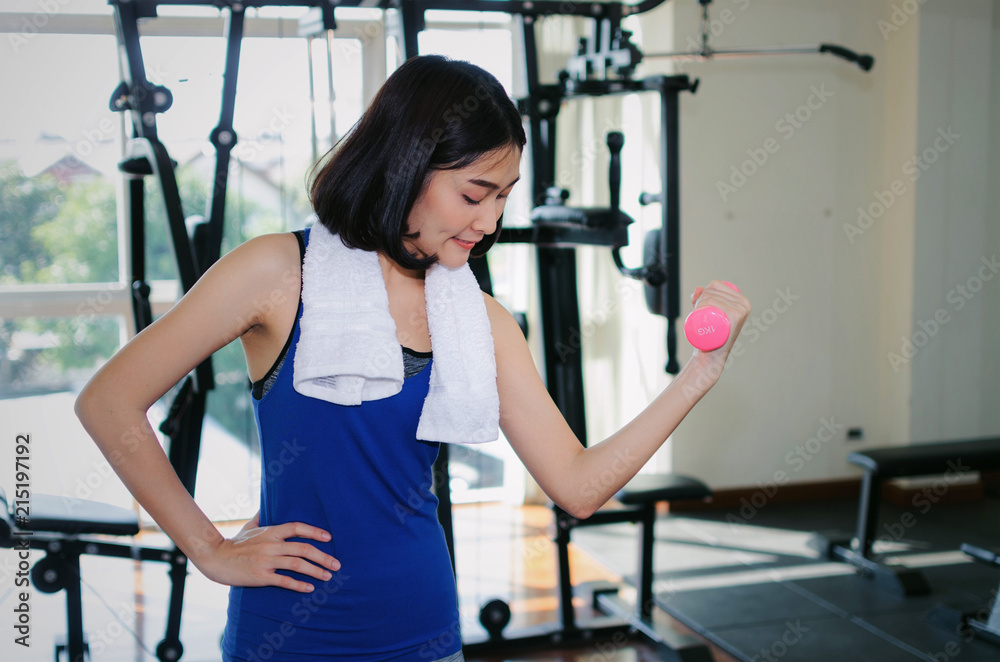 young asian pretty woman slim body doing exercises with dumbbell at biceps for good healthy in fitness gym at morning with weight machine background, bodybuilder, lifestyle and sport exercise concept