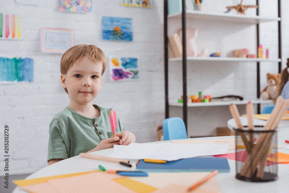portrait of preschooler boy sitting at table with paper and colorful pencils for drawing in classroom