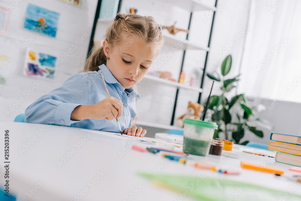 portrait of adorable focused child drawing picture with paints and brush at table