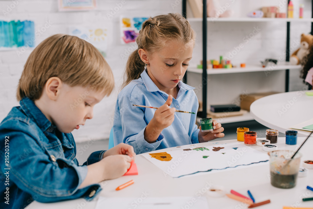 Fototapeta premium focused preschoolers drawing pictures with paints and paint brushes at table in classroom
