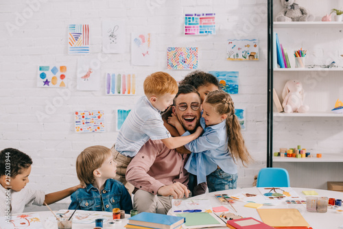 Obraz na plátně interracial kids hugging happy teacher at table in classroom
