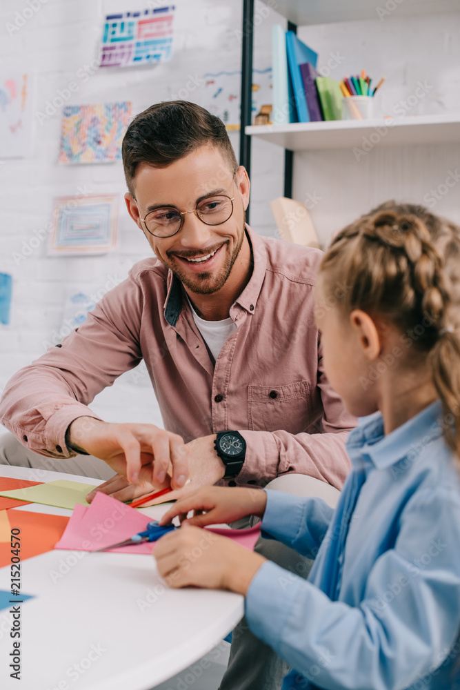 Obraz premium smiling teacher and cute preschooler cutting papers with scissors att able in classroom