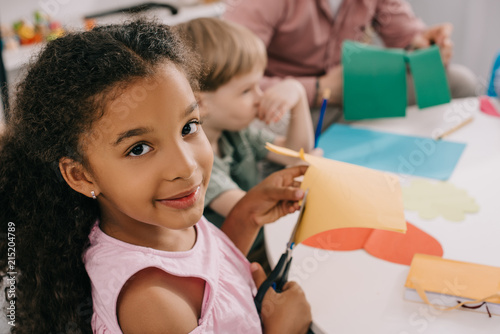 selective focus of multiethnic preschoolers cutting colorful papers with scissors in classroom photo
