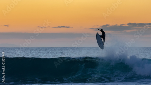 The silhouette of a surfer in the air above a wave at sunrise with a golden sky in the background