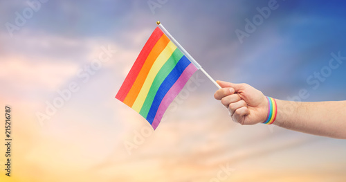 lgbt  same-sex relationships and homosexual concept - close up of male hand with gay pride awareness wristband holding rainbow flag over evening sky background