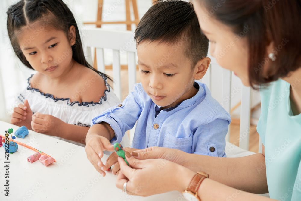 © DragonImages - Teacher woman showing and helping with plasticine to cute Asian boy and girl in art class