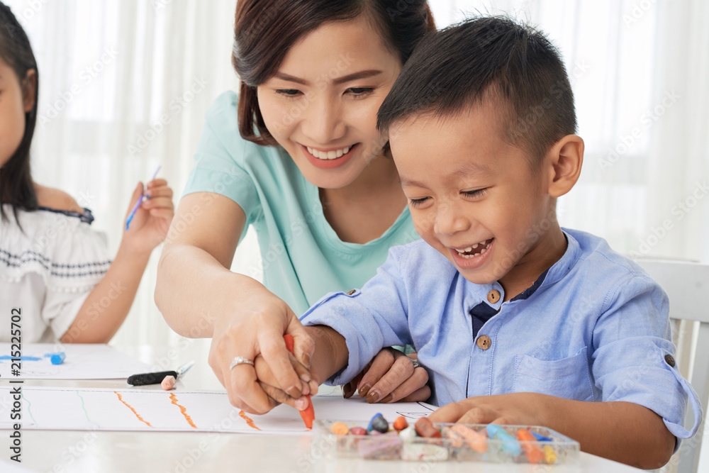 Smiling Asian boy drawing with teacher and having fun in art class