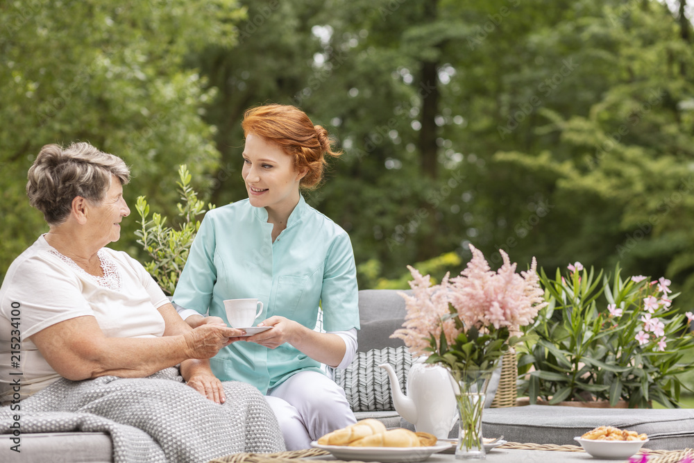 Happy nurse giving tea to elderly woman while eating breakfast on the terrace Happy nurse giving tea to elderly woman while eating breakfast on the terrace
