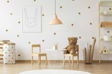 Teddy bear on wooden chair next to table in gold kid's room interior with lamp and poster. Real photo