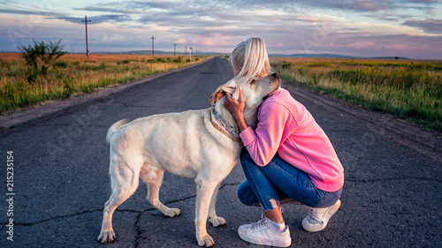 blonde girl hugging a white Labrador dog on the road in a beautiful dawn