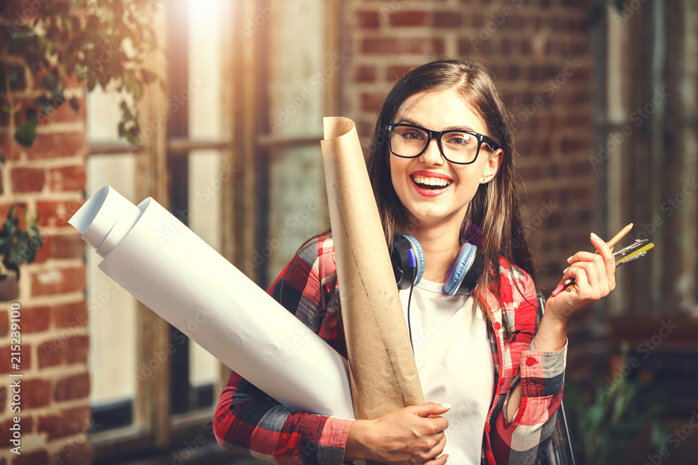 Cute young long-hair female architect wearing glasses, with headphones holding blueprints and smiling near the window in the loft modern office Cute young long-hair female architect wearing glasses, with headphones holding blueprints and smiling near the window in the loft modern office