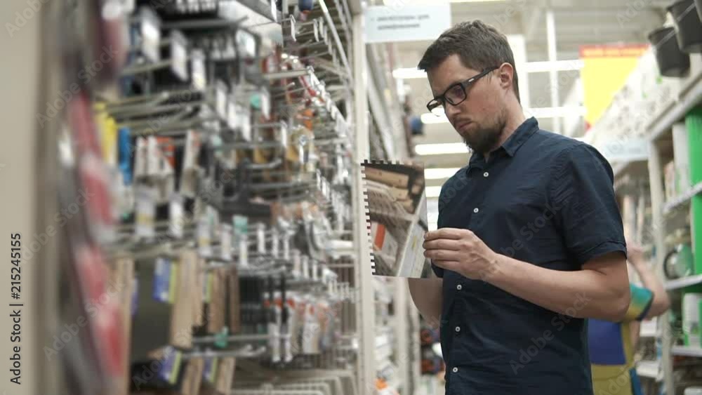 Master is choosing tool for a finishing works and tiling in a shop for builders. Man is looking on a toothed spatula, standing near rack Master is choosing tool for a finishing works and tiling in a shop for builders. Man is looking on a toothed spatula, standing near rack