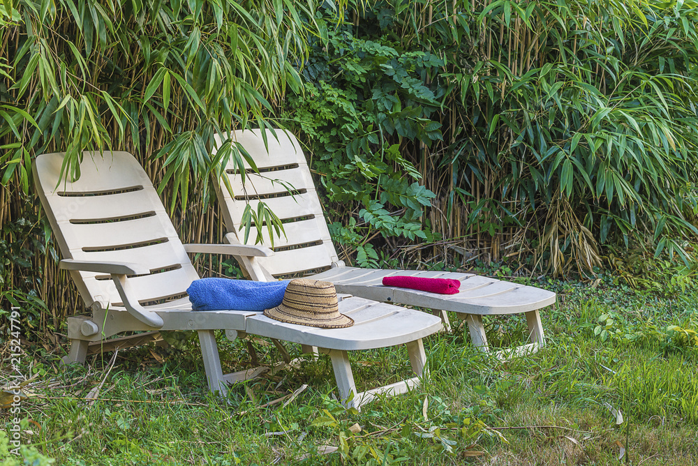 Holiday concept: two white lounge chairs, towels, straw hat in greenery of bamboo