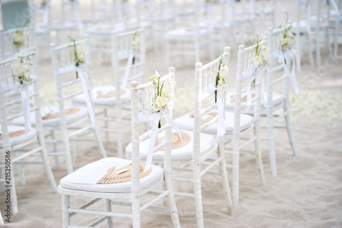 Side view of white chiavari chairs, wedding chairs arrangement for beach wedding venue on the white sand outdoors, White ribbin with rose flower flower deoration