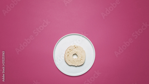 Half plain bagel on a white plate on a pastel pink background