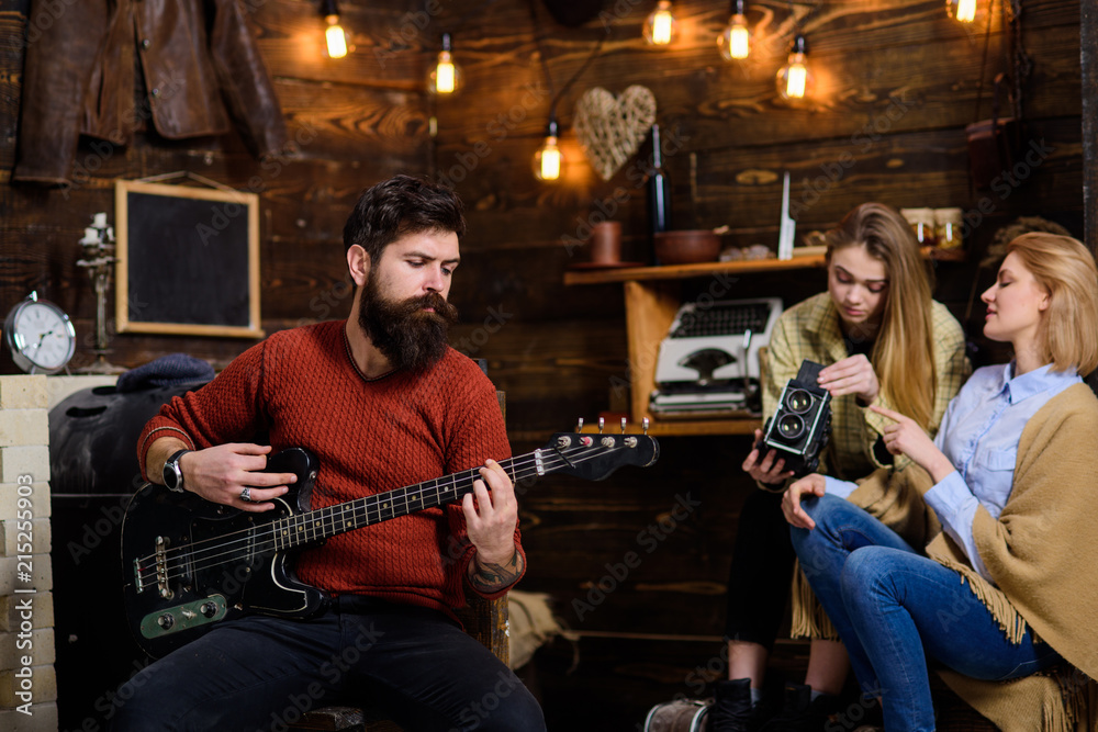Obraz premium Man with long beard enjoying music. Musician with calm, concentrated face playing for his wife and daughter. Mom helping teenage girl to deal with retro camera. Bearded man tuning electrical guitar