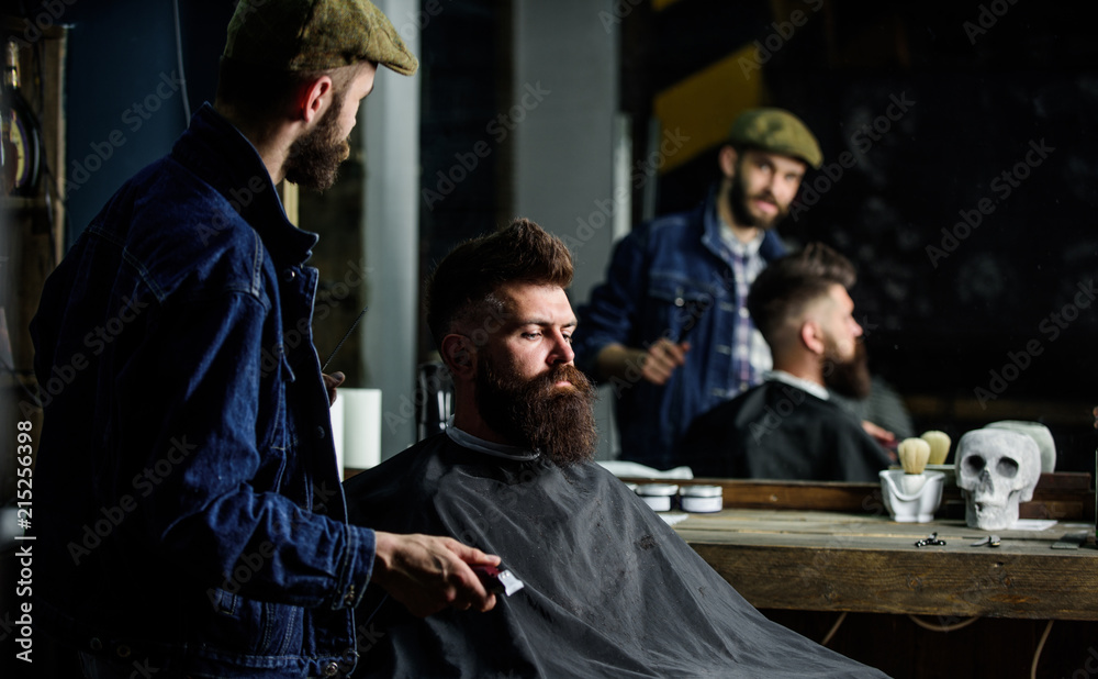 Obraz premium Hipster client got new haircut. Barber with hair clipper looking at mirror, barbershop background. Professional master checking result while client sits in chair. Barber done haircut. Haircut concept