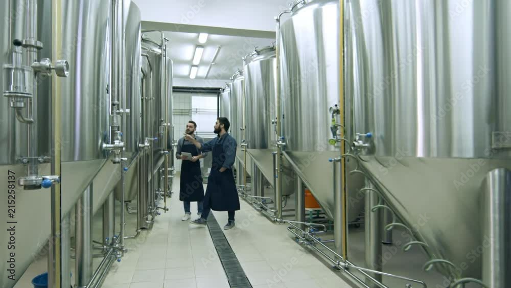 Two coworkers walking through beer factory between rows of steel brewing vessels, using digital tablet, inspecting equipment and talking Two coworkers walking through beer factory between rows of steel brewing vessels, using digital tablet, inspecting equipment and talking