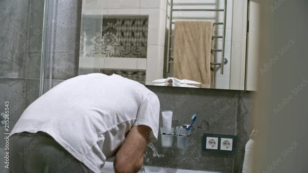 Rear view of mature man in white t-shirt washing face with tap water and looking in the mirror when getting ready for work in the morning Rear view of mature man in white t-shirt washing face with tap water and looking in the mirror when getting ready for work in the morning