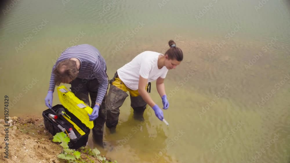 Man and woman scientist environmentalist standing in a river. Woman taking sample of water and pouring it into the test tube. Man and woman scientist environmentalist standing in a river. Woman taking sample of water and pouring it into the test tube.