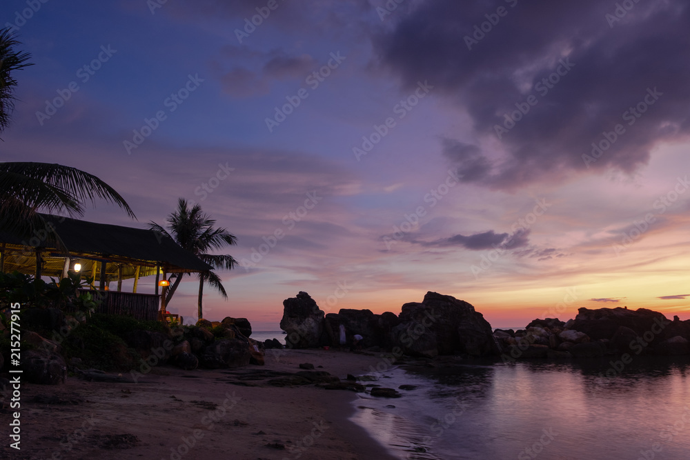 Beach, rocks and beach bar shadowed under the palm trees on the background of the sunset almost dark sky with vibrant orange, blue and purple colours