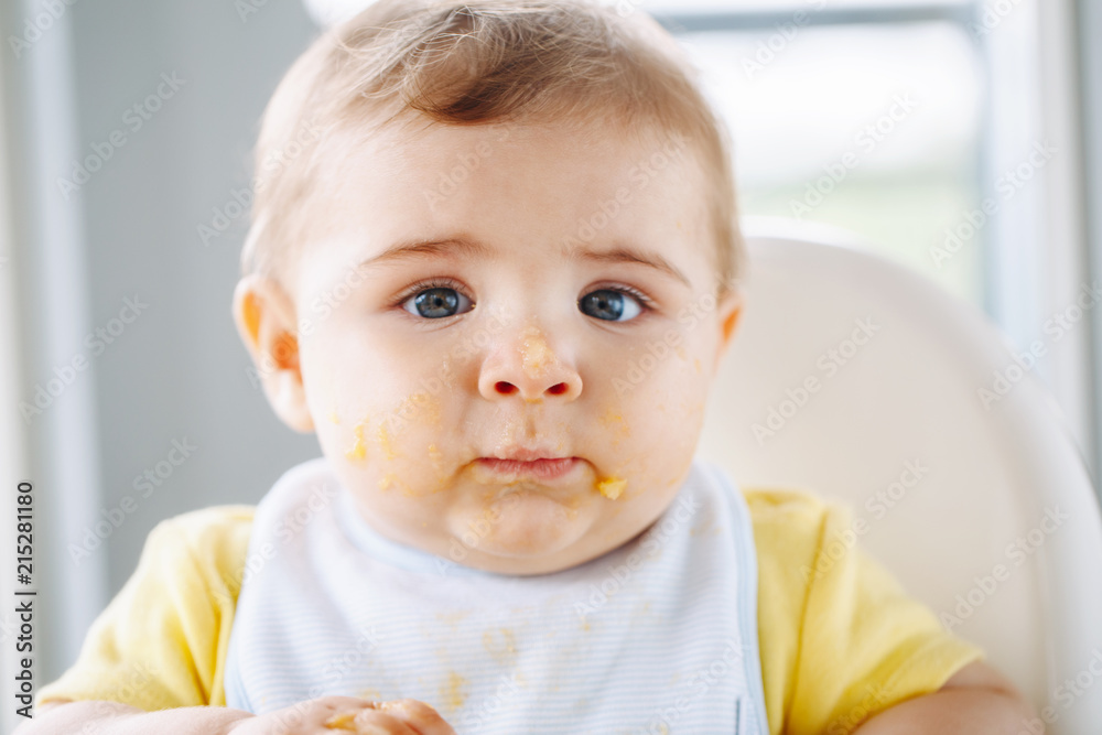 Portrait of cute adorable Caucasian child boy with dirty messy face sitting in high chair eating apple puree with fingers. Everyday home childhood lifestyle. Infant trying supplementary baby food Portrait of cute adorable Caucasian child boy with dirty messy face sitting in high chair eating apple puree with fingers. Everyday home childhood lifestyle. Infant trying supplementary baby food