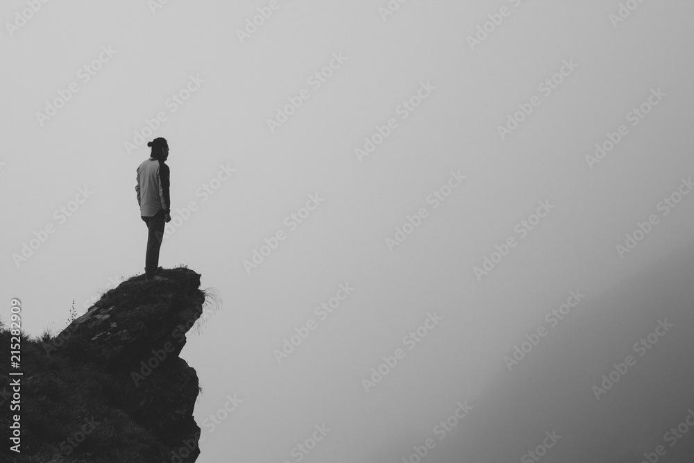 Black and white image of a man standing at the edge of the cliff against a  foggy landscape. Photos