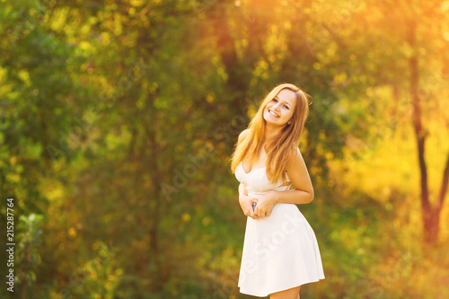 Stylish white girl dress who holding phone in her hands and beautifully smiling against the background of green trees