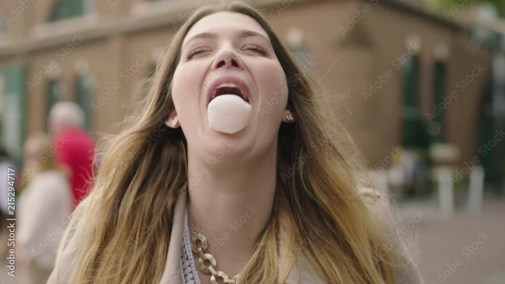 portrait of young happy blonde woman blowing bubblegum laughing optimistic on urban background