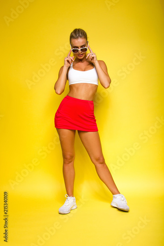 young sports happy sexy blonde girl in red skirt and white top is posing  like model and looking down with sun glasses in her hands on the yellow  wall background Stock Photo
