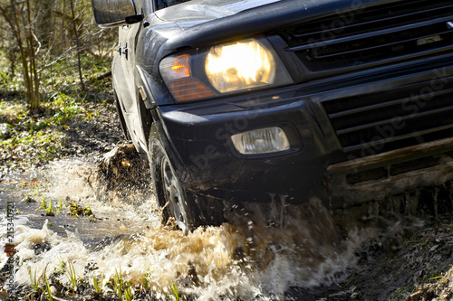SUV close-up front of it passes over rough terrain, through a puddle on a dirty road. Sharpness in the middle field