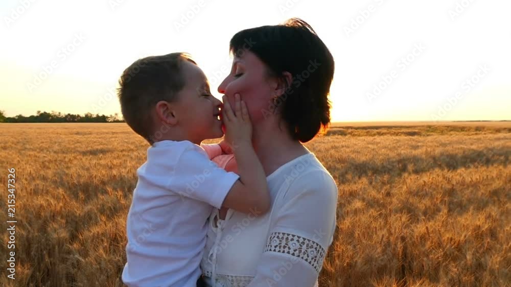 A happy mother holds a child in her arms in a field of wheat against a sunset background. The child kisses the mother's face, the mother kisses the child A happy mother holds a child in her arms in a field of wheat against a sunset background. The child kisses the mother's face, the mother kisses the child