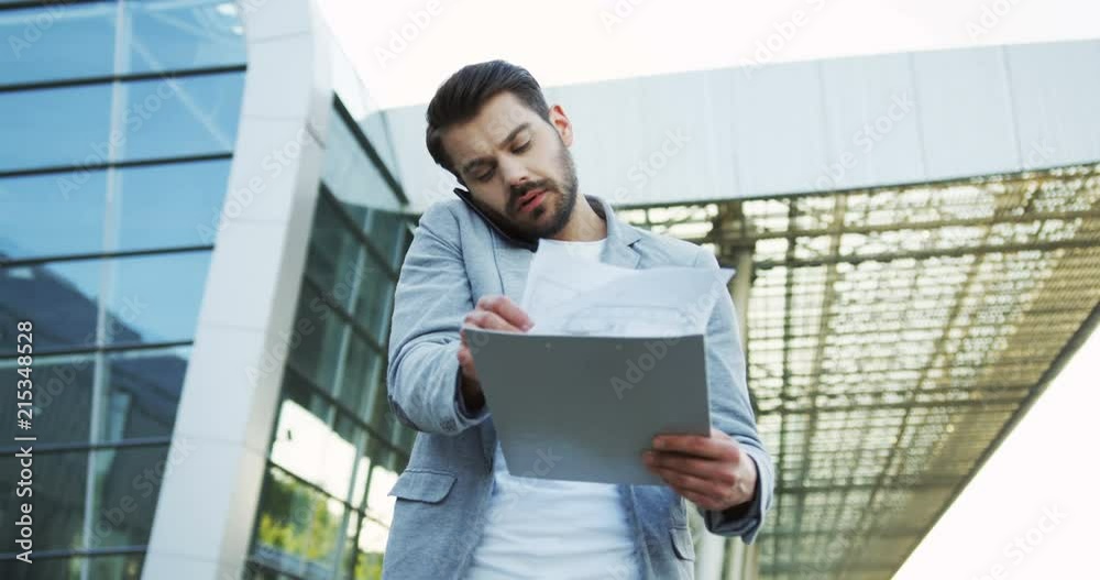 Close up of the caucasian businessman walking with some documents in hands which he checking while rushing to the office and talking on the phone. Close up of the caucasian businessman walking with some documents in hands which he checking while rushing to the office and talking on the phone.