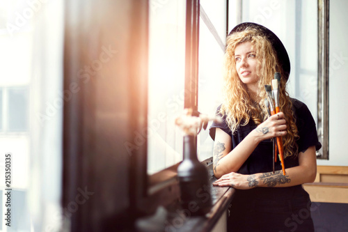 Blonde long hair female artist in black clothes looking in the window, she smiling and holding her brush