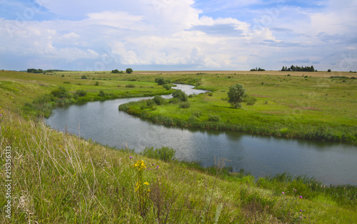 Sunny summer landscape with small river flowing between the green hills and fields.Beautiful dark clouds in blue sky.