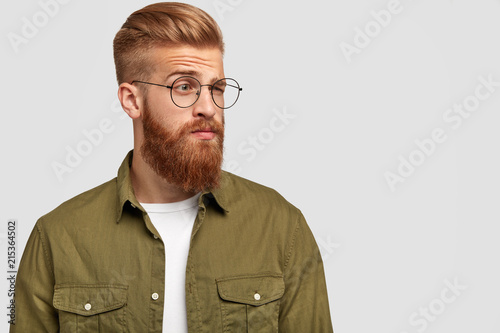 Indignant ginger male looks aside with puzzled thoughtful expression, raises eyebrows, wears glasses, wears fashionable clothing, stands against white background with copy space for your promotion photo