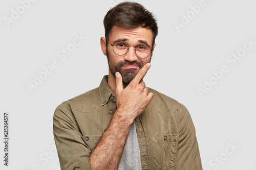 Indoor shot of puzzled hesitant unshaven guy holds chin and doubts, raises eyebrows, has clueless expression, wears stylish shirt, isolated over white background. People, emotions, lifestyle concept photo