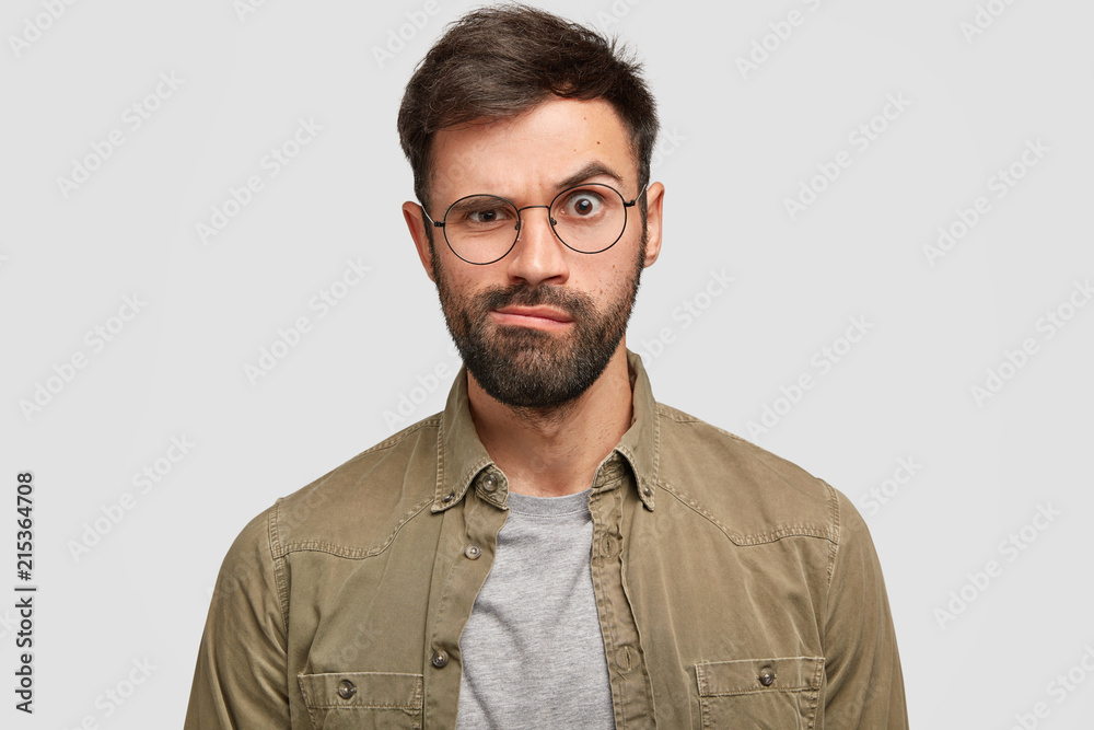 Displeased angry European man raises eyebrows and purses lips furiously, dressed in fashionable shirt, expresses negative emotions, isolated over white background. Facial expressions concept