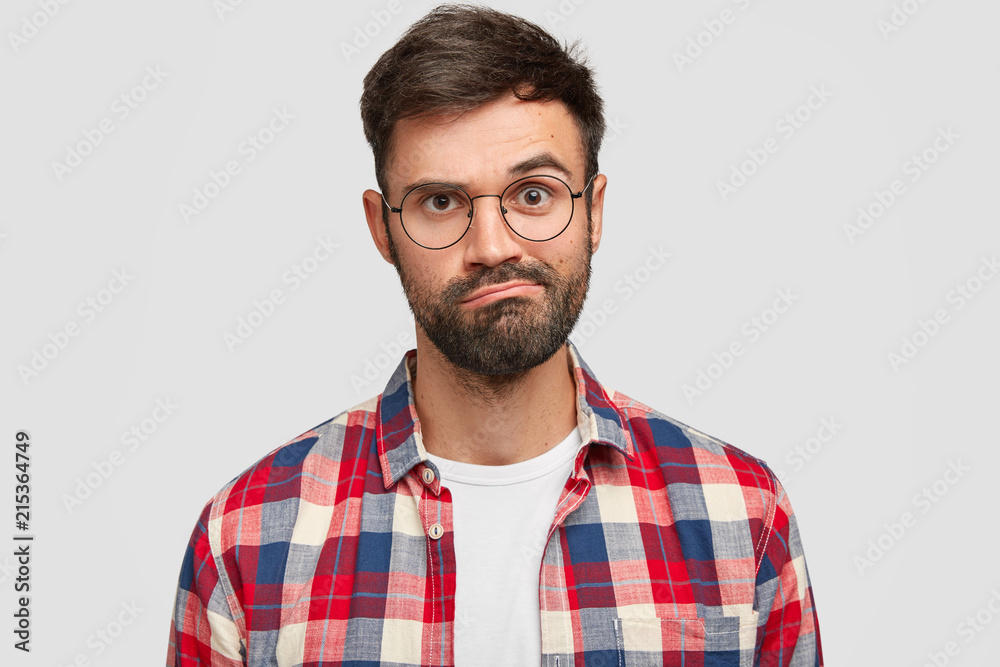 Fototapeta premium Headshot of puzzled hesitant displeased unshaven young guy purses lips, has uncertain exression, trendy haircut, wears checkered shirt, isolated over white background. Facial expressions concept