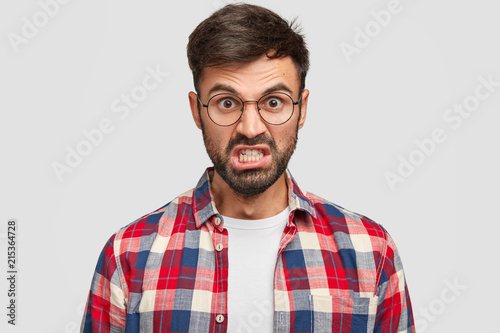 Photo of irritated unshaven man looks angrily at camera, clenches teeth and raises eyebrows, being annoyed with much duties on work, dressed in checkered shirt, stands against white background. © Wayhome Studio